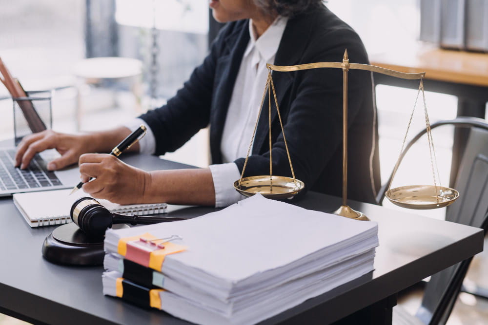 Gros plan sur une avocate en droit du travail à son bureau, signant des documents et utilisant un ordinateur portable, avec une balance de justice en laiton et un marteau à proximité